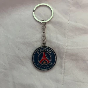 Nyckelring med Paris Saint-Germain logo - Säljer en rund nyckelring med Paris Saint-Germain fotbollsklubbens emblem. Nyckelringen är i metall med blå, röd och vit färg och har klubbens klassiska logga framtill. Perfekt för dig som är PSG-supporter!
