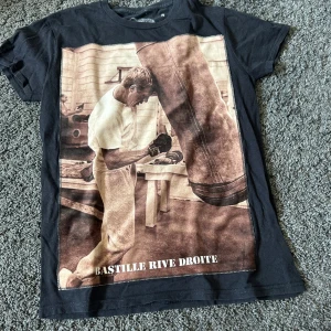 Svart t-shirt med boxningsmotiv från Bastille - Cool svart t-shirt från Bastille med stort tryck framtill av en boxare och boxningssäck. Texten 'BASTILLE RIVE DROITE' syns på framsidan. Tillverkad i 100% bomull och gjord i Italien. Perfekt för dig som gillar sportiga och unika plagg. 