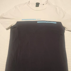 Hugo boss tröja  - Snygg t-shirt från BOSS i en stilren design med vit överdel och svart underdel. Den har korta ärmar och ett subtilt blått tryck på bröstet.