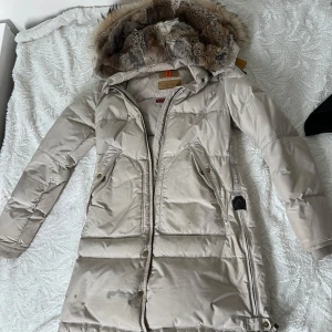 Ljusbeige dunjacka med päls från Parajumpers - Säljer en lång, ljusbeige vinterjacka från Parajumper. Jag säljer på grund av att den är för liten för mig. Den har en liten fläck på nederdelen av jackan som man kan se på bilden men annars ser den helt ny ut.