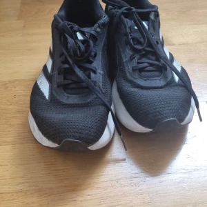 Svarta Adidas sneakers med vita detaljer - Säljer ett par svarta Adidas sneakers med vita ränder på sidorna och vit sula. Skorna har snörning och är tillverkade i syntetmaterial med mesh för bra andningsförmåga. Perfekta för träning eller vardagsbruk.