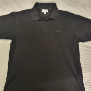 Lacoste pikétröja - Säljer en klassisk svart pikétröja från Lacoste med korta ärmar och den ikoniska krokodilloggan på bröstet. Tröjan har en krage och knappar framtill.