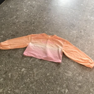 Croppad sweatshirt i pastell från H&M - Säljer en croppad sweatshirt från H&M i pastellfärger med texten 'FRIDAY FEELING' på bröstet. Tröjan har långa ärmar och en mjuk bomullskänsla. Färgerna går i en gradient från persika till ljusrosa.