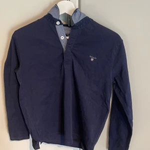 Mörkblå långärmad tröja från GANT - Säljer en stilren mörkblå långärmad tröja från GANT med krage och knappdetalj vid halsen. Tröjan har en klassisk passform och diskret logga på bröstet. Perfekt för dig som gillar en enkel men snygg look.Passar bra över skjortan