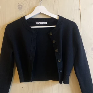 Svart kofta från Zara - Säljer en svart, croppad kofta från Zara med rund halsringning och stora knappar framtill. Tröjan har långa ärmar och är perfekt att styla över en topp eller klänning. Enkel och stilren design som passar till mycket.
