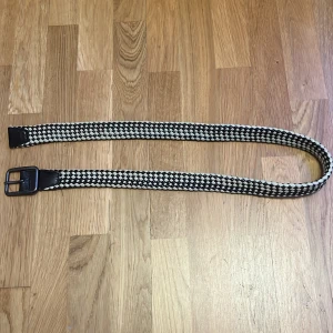 Flätat bälte från Armani  - Snyggt ”grisch” bälte från Armani Jeans med ett svart och beige rutigt mönster. Tillverkat i 100% bomull och läderdetaljer. Perfekt för att ge din outfit en stilfull touch.