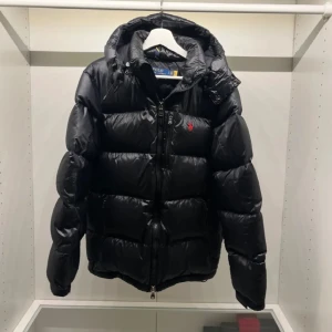 Svart dunjacka från Polo Ralph Lauren - Säljer en svart dunjacka från Polo Ralph Lauren med röd broderad logga på bröstet. Jackan har dragkedja framtill, hög krage och huva. Klassisk pufferstil med quiltade sektioner och ribbade muddar vid ärmslut. Skicket är 9/10 förutom en liten skada på ärmen så är den i jätte fint skick! Hör av er vid funderingar. Pris kan diskuteras vid snabb affär!😄