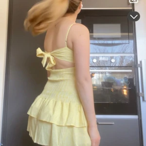 Gul rutig volangklänning med rosett i ryggen - Söt gul kortklänning med rutigt mönster och volangkjol. Klänningen har smala axelband, rosett i ryggen och omlott-detalj baktill. 💛