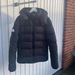 Vinterjacka Superdry - Säljer en svart pufferjacka från Superdry i bra skick. Jackan har en dragkedja framtill och en rymlig huva som går att ta av. Perfekt för kalla vinterdagar, aldrig haft problem med värme i denna. Det finns en liten reva på ena ärmen, går att fixa genom att sy ihop eller med en fix lapp men jag är ej kunnig inom det området. Tror att den är slutsåld online då jag ej hittar den. Köptes för ≈1300kr, hör av dig om du har minsta lilla fråga. Pris går att diskutera!