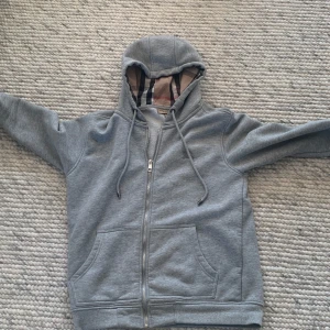 Grå hoodie från Burberry - Säljer en stilren grå hoodie från Burberry med dragkedja och klassiskt rutigt mönster i luvan. Perfekt för en avslappnad och trendig look. Hoodien har långa ärmar och praktiska fickor framtill. STORLEK M! köpte denna hoodien för en 1 månad sen så den är Ny