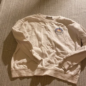Vit sweatshirt med Hawaii-tryck - Säljer en vit sweatshirt med tryck på bröstet där det står 'Hawaii Surfing Island The Perfect Vibe' och en regnbåge ovanför. Tröjan har långa ärmar och ribbade muddar vid ärmslut och nederkant. Perfekt för en avslappnad stil.