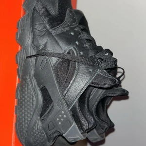 Svarta Nike Air Huarache sneakers - Säljer ett par helsvarta Nike Air Huarache sneakers med klassisk chunky sula och snörning. Skorna har en mix av skinn och syntetmaterial, vilket ger en cool och sportig look. Perfekta för dig som gillar streetwear och vill ha något bekvämt och stilrent.