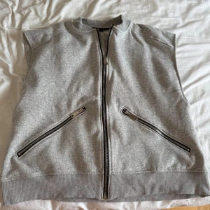 Grå väst med dragkedjor - Snygg grå väst med ribbad krage och nederkant. Västen har en rak passform och är försedd med dragkedja framtill samt två snedställda fickor med dragkedjor. Perfekt att slänga över en hoodie eller t-shirt för en avslappnad look.