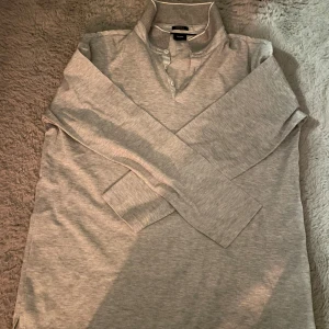 Ljusgrå pikétröja från Hugo Boss - Säljer en stilren ljusgrå pikétröja från Hugo Boss i storlek L. Tröjan har lång ärm, klassisk krage och knappar vid halsen. Tillverkad i bomull med merceriserad finish för extra mjuk känsla och snygg lyster.