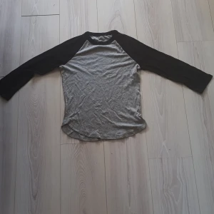 Grå och svart långarmad tröja - Säljer en grå och svart raglantröja med långa ärmar. Perfekt för en avslappnad look. Tröjan har en rund halsringning och är tillverkad i ett mjukt material.