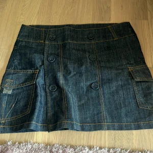 Jeanskjol från Clara - Snygg jeanskjol från Clara med knappar framtill och praktiska fickor på sidorna. Kjolen har en kort längd och är perfekt för en avslappnad stil. Den mörkblå denimfärgen ger en klassisk look.