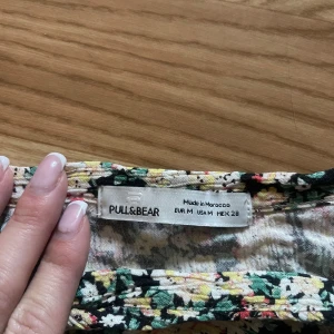 Blommig omlottopp från Pull&Bear - Supersöt croppad omlottopp från Pull&Bear med blommigt mönster i grönt, vitt, beige och rosa. Toppen har volangdetaljer vid axlarna och v-ringning, samt smockad nederkant för en snygg passform. Perfekt för vår och sommar!