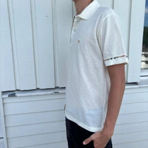 Burberry pike  - Snygg vit kortärmad pikétröja från Burberry med klassiska rutiga detaljer vid knäppningen och ärmsluten. Tröjan har en liten broderad logga på bröstet och är tillverkad i mjuk bomull. Perfekt för dig som vill ha en clean och stilren look med lyxig känsla.