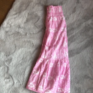 Rosa långkjol med vitt mönster - Supersöt rosa långkjol med vitt mönster och smock i midjan. Kjolen har en flowy passform och volang nertill som ger en bohemisk vibe. Perfekt för varma dagar och lätt att styla med en enkel topp. Materialet känns mjukt och luftigt. Storlek är 146-152