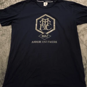 Tottenham Hotspur Blå t-shirt - Snygg svart t-shirt från Tottenham Hotspur med stort tryck framtill i silver med klubbens klassiska emblem och texten 'Audere Est Facere 1882'. Skön passform och rund halsringning, perfekt för dig som älskar fotboll och vill visa ditt lag.