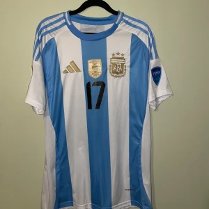 Argentina Garnacho #17 Copa America tröja - Argentina landslagströja från Adidas med Garnacho och nummer 17 på ryggen. Klassiska blå och vita ränder, guldiga mästerskapsmärken och Copa America 2024-patch på ärmen. Tillverkad i lätt och ventilerande material med Aeroready-teknologi. Perfekt för dig som älskar fotboll och landslagskänsla.