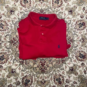 Röd pikétröja från Polo Ralph Lauren - Klassisk röd pikétröja från Polo Ralph Lauren med blå broderad logga på bröstet. Tröjan har korta ärmar, krage och två knappar framtill. Tillverkad i mjuk bomull som är skön mot huden. Perfekt för en clean och sportig look.