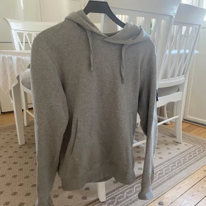 Grå hoodie från Stone Island - Säljer en grå hoodie från Stone Island med klassisk logga på ärmen. Tröjan har huva med snörning, magficka och långa ärmar. Perfekt för dig som gillar streetwear och vill ha en stilren look. Materialet är mjuk bomull för skön känsla.