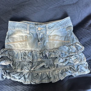 Jeanskjol med volanger - Supersöt ljusblå jeanskjol med flera lager volanger och fickor fram. Kjolen har bälteshällor, knapp och dragkedja framtill. Perfekt för dig som vill ha en trendig och lekfull look.