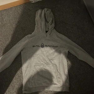 Grå hoodie från Sail Racing - Snygg ljusgrå hoodie från Sail Racing med svart logga på bröstet och en mindre logga på huvan. Klassisk känguruficka framtill och mjukt material som känns skönt mot huden. Perfekt för dig som gillar en clean och sportig stil.