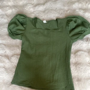 Grön topp med puffärmar - Supersöt grön topp med fyrkantig ringning och puffiga korta ärmar. Tyget har en diskret struktur som ger en cool känsla. Perfekt att styla med jeans eller kjol för en trendig look. Färgen är en härlig grön som passar året runt.
