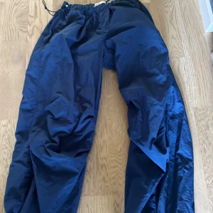 Blå täckbyxor från Bershka - Mörkblå parachute byxor från Bershka med elastisk midja och dragsko. Byxorna är tillverkade i ett syntetmaterial som känns lätt och smidigt. Perfekta för kalla dagar när du vill hålla dig varm och torr. Snygg loose passform och enkel att dra på.