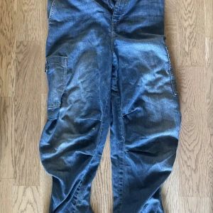 Blå cargopants från Divided, M - Säljer ett par blå cargopants från Divided i jeansmaterial. Byxorna har elastisk midja, stora sidofickor och klassisk cargostil. Perfekta för en avslappnad och cool look. Passar dig som gillar streetwear och praktiska detaljer.