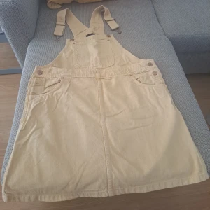 Beige hängselklänning i bomull - Supersöt beige hängselklänning i mjuk bomull med klassiska fickor fram och justerbara axelband. Perfekt för lager-på-lager och enkel att styla med t-shirt eller tröja under. Rak passform och praktiska knappar i sidorna.