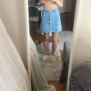 Jeanskjol med knappar - Säljer en ljusblå jeanskjol från H&M Divided i storlek 34. Kjolen är kort, har en A-linjeform och snygga silverfärgade knappar framtill. Klassisk denim med bälteshällor och sömmar som ger en cool vintagekänsla. Perfekt till sneakers eller boots!