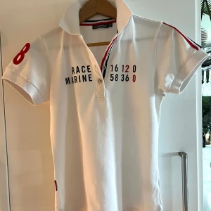 Vit piké från Race Marine, storlek 36 - Vit pikétröja från Race Marine med korta ärmar, broderade siffror på ärmarna och tryck framtill. Klassisk krage och knäppning med tre knappar. Röd och marinblå detalj längs insidan av knappslån. Skön bomullskvalitet, perfekt för en sportig look. Den är i mycket bra skick, som en ny. Storleken kan man se på den första bilden 