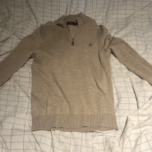 Beige merinoullströja från Morris - Snygg beige stickad tröja från Morris i 100% merinoull. Tröjan har half zip med läderdetalj, ribbade muddar och diskret broderad logga på bröstet. Perfekt för lager-på-lager och stilren look.