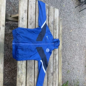 Blå Nike PSG vindjacka M - Snygg blå vindjacka från Nike med PSG-logga på bröstet. Jackan har svarta och vita detaljer, hel dragkedja och huva. Perfekt för dig som gillar sportig stil och vill sticka ut med en ikonisk klubbjacka.