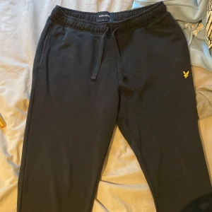 Svarta mjukisbyxor från Lyle & Scott - Svarta mjukisbyxor från Lyle & Scott med resår och snörning i midjan. Byxorna har en gul broderad logga på vänster höft och är tillverkade i mjuk bomull. Perfekta för chill eller träning.