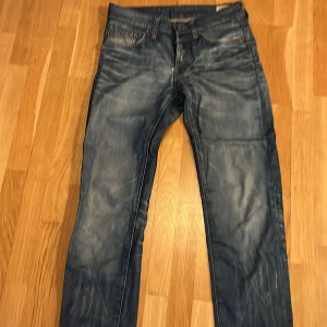 Blå jeans från G-Star RAW - Säljer ett par snygga blå jeans från G-Star RAW med coola slitningar och detaljerade sömmar. Jeansen har klassisk femficksdesign, läderpatch bak och dragkedjor på bakfickorna. Perfekta för dig som gillar streetwear och en avslappnad look.