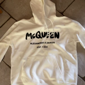 Vit hoodie från Alexander McQueen - Säljer en vit hoodie från Alexander McQueen med svart tryck framtill. Tröjan har huva med snörning, stor magficka och långa ärmar. Perfekt för dig som gillar streetstyle och vill ha något snyggt och bekvämt med ikonisk logga.
