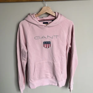 Ljusrosa hoodie från GANT med logga - Säljer en ljusrosa hoodie från GANT med broderad logga och sköld på bröstet. Tröjan har huva, känguruficka och långa ärmar. Materialet är mjuk bomullsmix som känns skön mot huden. 