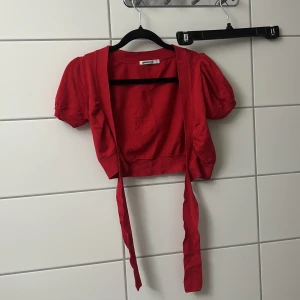 Röd bolerotopp med knyt från Gina Tricot - Kort röd bolerotopp från Gina Tricot med puffiga korta ärmar och långa knytband framtill. Perfekt för att lägga till en färgklick till din outfit. Toppens material känns mjukt och stretchigt.