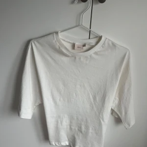 Vit croppad topp med knyt från Nelly - Säljer en vit croppad topp från Nelly med rund halsringning och korta ärmar. Toppen har en snygg knytning i ryggen som ger en cool detalj. Perfekt till jeans eller kjol för en clean look.