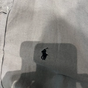 Grå skjorta från Ralph Lauren - Snygg grå skjorta från Ralph Lauren med klassisk button-down krage och broderad logga på bröstet. Skjortans har lång ärm, knäppning framtill och är tillverkad i bomull för en skön känsla. Perfekt för dig som gillar stilrena och tidlösa plagg.