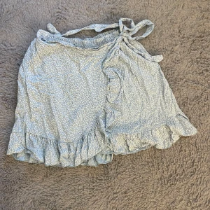 Ljusblå omlottkjol med volang - Supersöt ljusblå kort omlottkjol med små vita blommor och volangkant. Kjolen har knyt i midjan och är perfekt för sommaren, köpt på en semester i spanien.