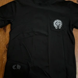  Svart Chrome Hearts t-shirt med tryck - Svart t-shirt från Chrome Hearts med stort vitt tryck på ryggen och mindre logga på bröstfickan. Snygga detaljer med CH-tryck nertill och klassisk passform. Perfekt för dig som gillar streetwear och vill sticka ut med en ikonisk design.