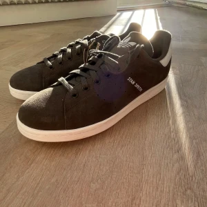 Adidas Stan Smith mocka sneakers - Snygga Adidas Stan Smith sneakers i mörkgrå mocka med vita detaljer och klassisk vit sula. Skorna har snörning, rund tå och ikoniska Adidas-loggor på plös och häl. Perfekta för dig som gillar stilrena och tidlösa sneakers. Tveka inte på att höra av dig!!