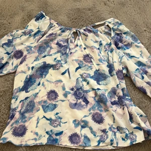Blommig blus från BikBok i blått - Supersöt blommig blus från BikBok i vitt och blått med lila toner. Blusen har långa ärmar, knytning vid halsen och ett luftigt, flowy material som känns perfekt för vår och sommar. Snygg att styla med jeans eller kjol för en fräsch look.