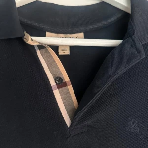 Mörkblå/navy pikétröja från Burberry  - Klassisk mörkblå/navy pikétröja från Burberry. Pikén har korta ärmar, krage och den ikoniska rutiga detaljen vid knäppningen. Broderad Burberry-logga på bröstet. Perfekt för en stilren och tidlös look. Ordinariepris 1600 kr.