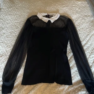 Svart blus med vit krage från Morgan de Toi - Supersnygg svart blus från Morgan med vit spetsig krage och transparenta långärmade ärmar i mesh med diskreta prickar. Kroppen är ribbad och har dragkedja i nacken. Perfekt för dig som gillar kontraster och coola detaljer.